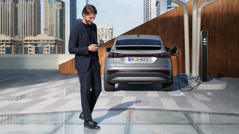 Ein Mann schaut auf sein Smartphone, während ein silberner Audi Q4 e-tron an einer Ladesäule in der Stadt aufgeladen wird.