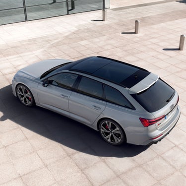 Silver car parked on a paved area near glass building and bollards, captured from above.