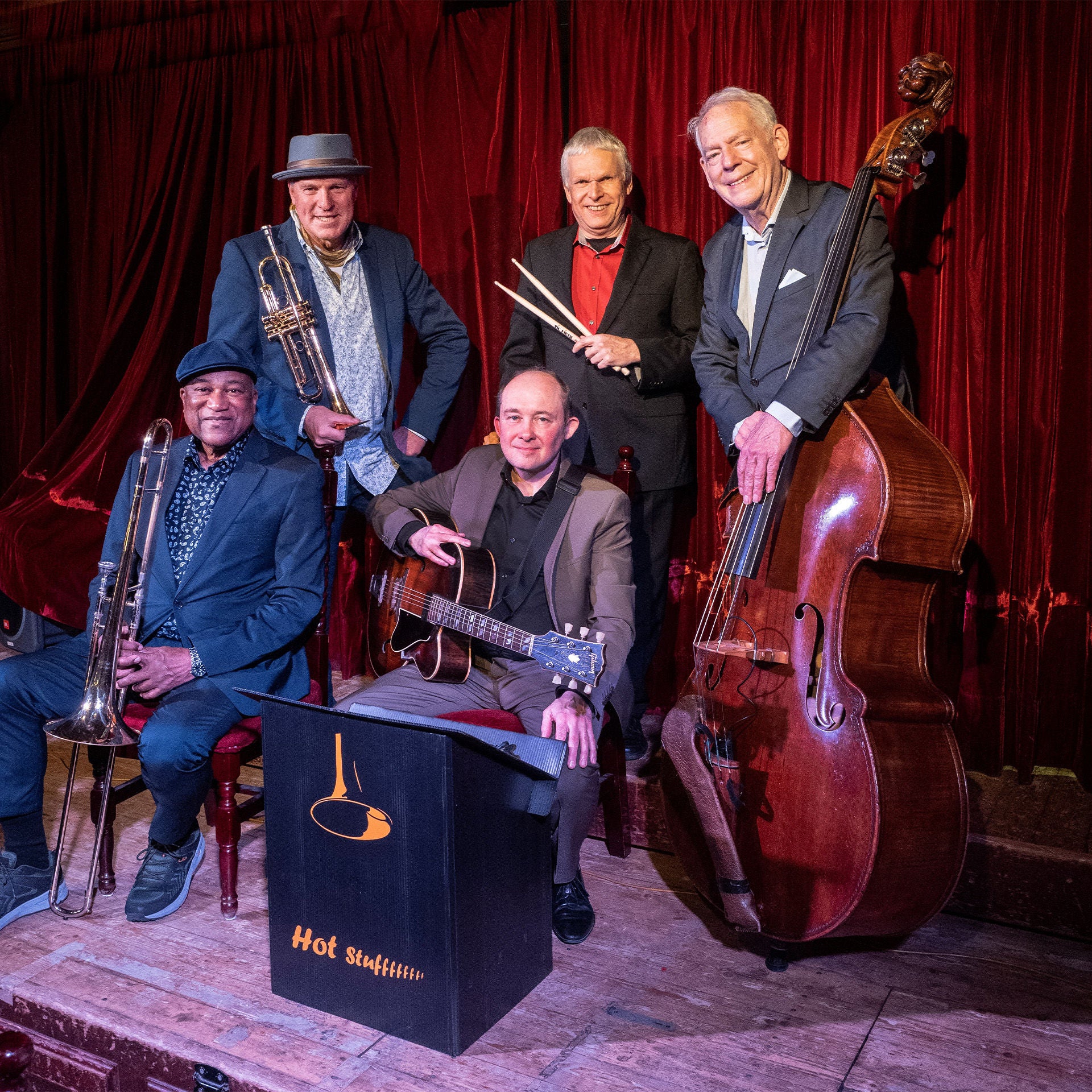 Fünf Musiker stehen und sitzen mit Instrumenten vor rotem Bühnenvorhang, darunter Kontrabass, Gitarre, Trompete und Schlagzeugstöcke.