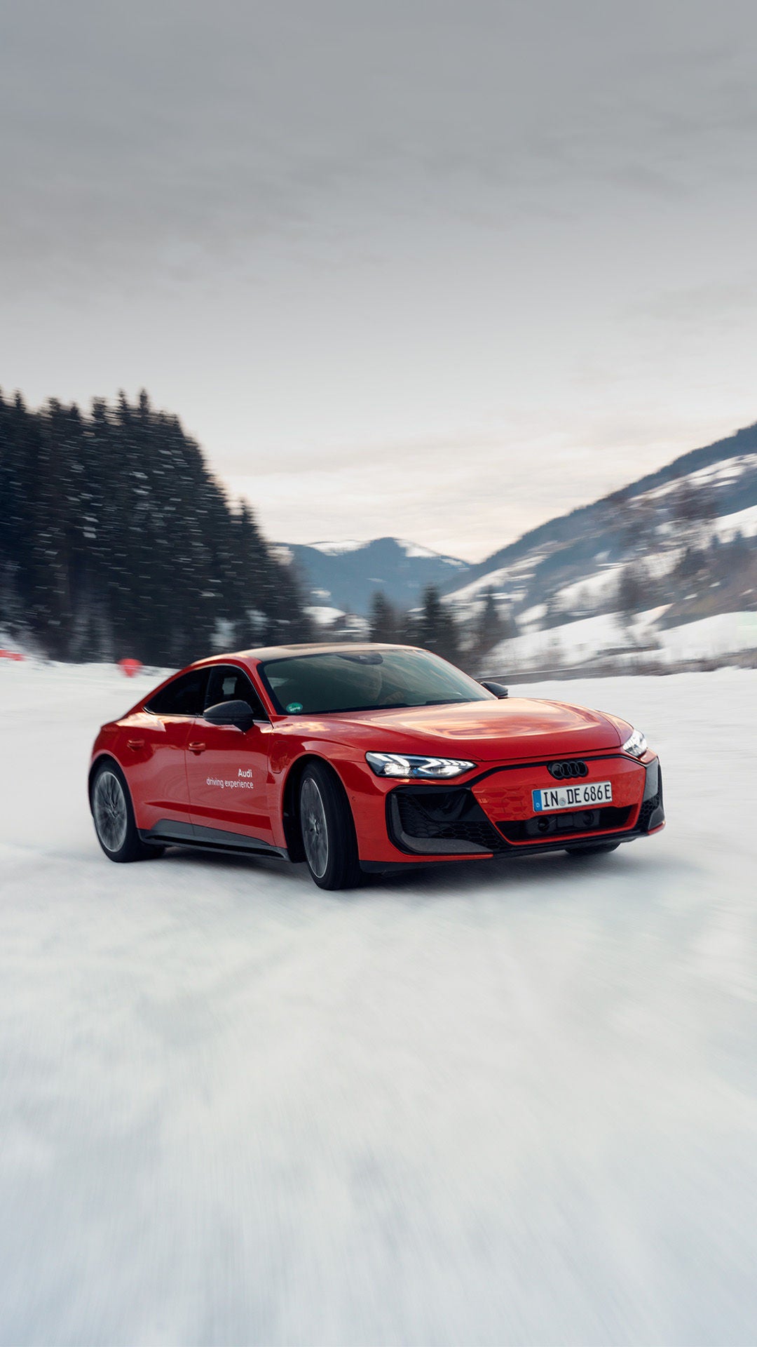 A red Audi RS e-tron GT performance speeds across a snow-covered area; snow dust, a forest, and mountains in the background.