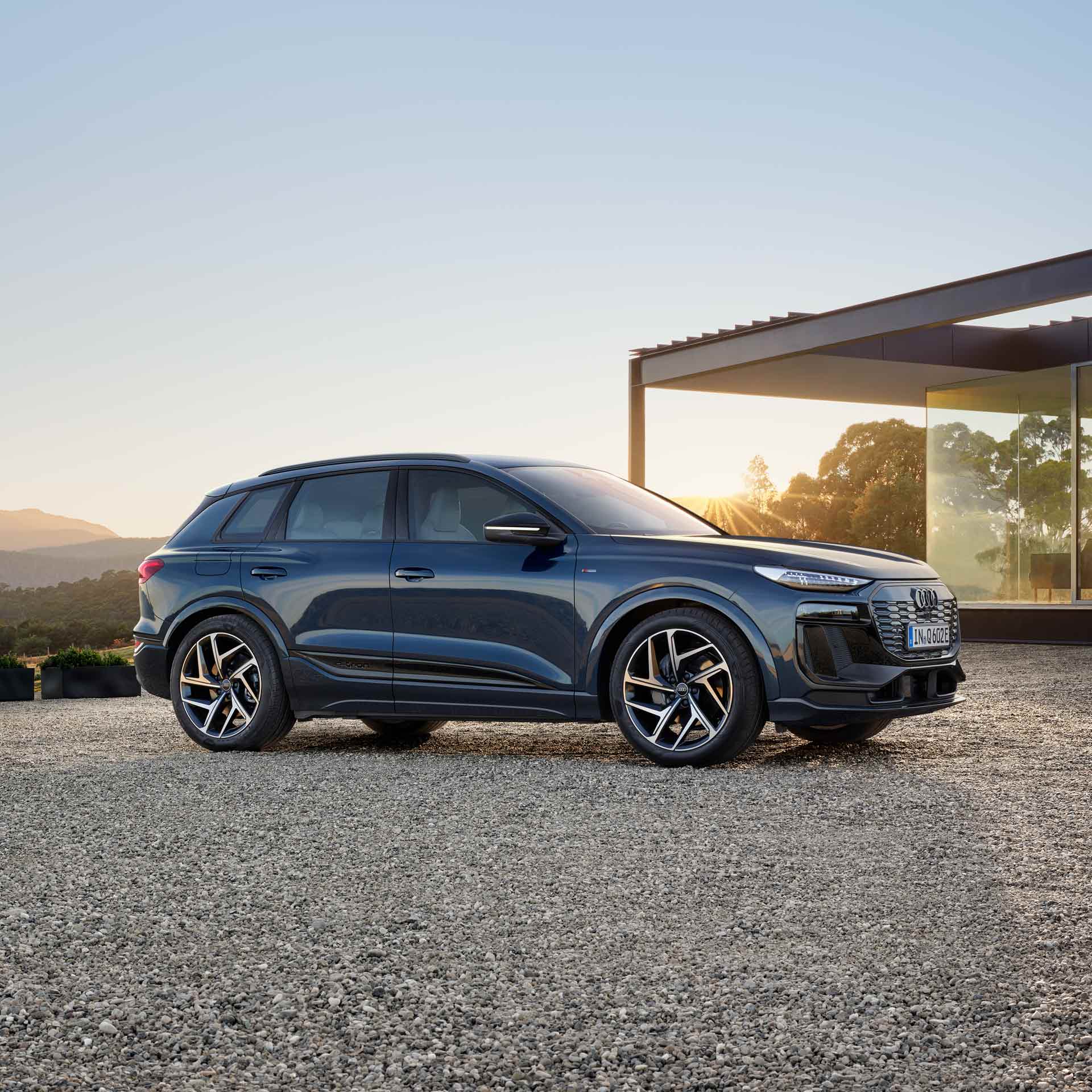 Ein blauer Audi Q6 SUV e-tron steht auf Kies vor einem modernen Haus, die Sonne scheint durch die Glasfassade, im Hintergrund eine bergige, grüne Landschaft.