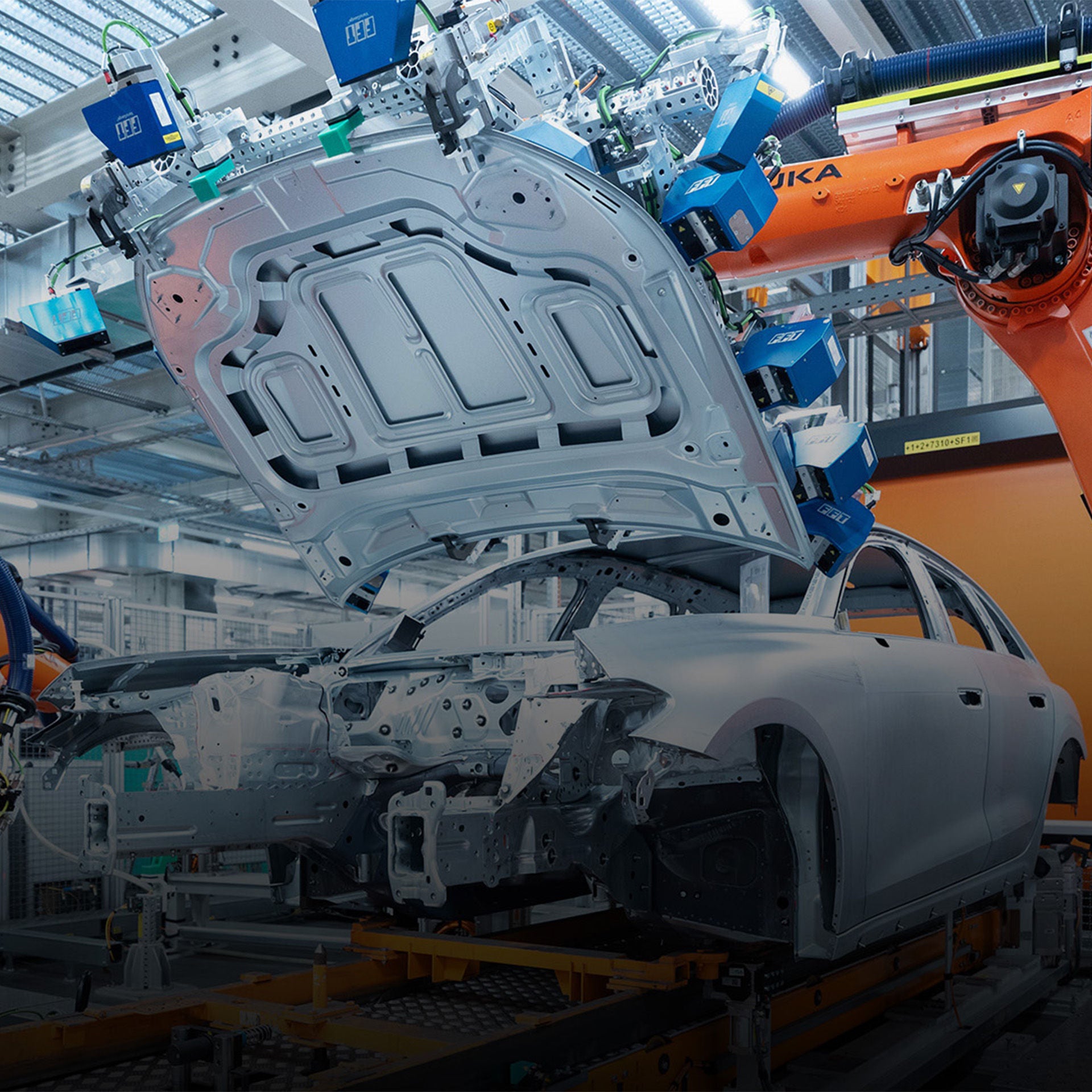 Unlackierte Autokarosserie mit geöffneter Motorhaube in einer Produktionshalle neben orangefarbenen Roboterarmen.