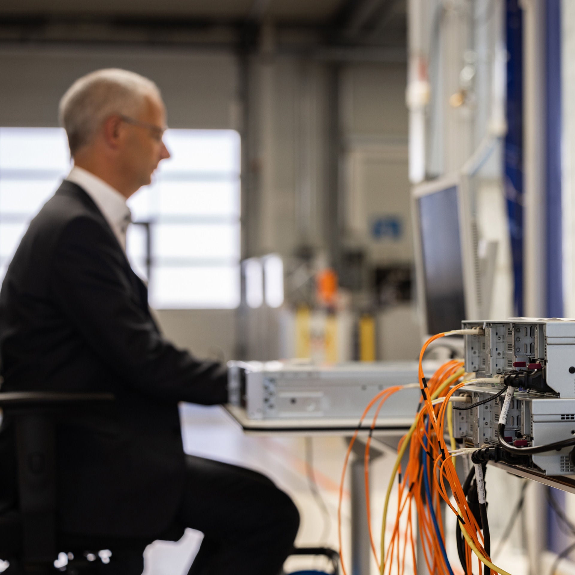 Bauteil, aus dem orangene Kabel herausragen. Im Hintergrund sitzt Henning Löser an einem Computerarbeitsplatz im P-Lab