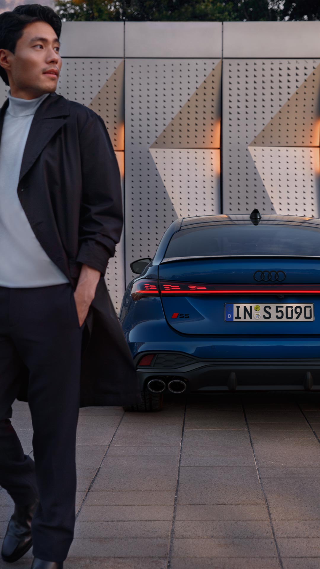 Blaue Audi S5 Limousine fährt auf einer Straße vor einer modernen Skyline bei Sonnenuntergang.