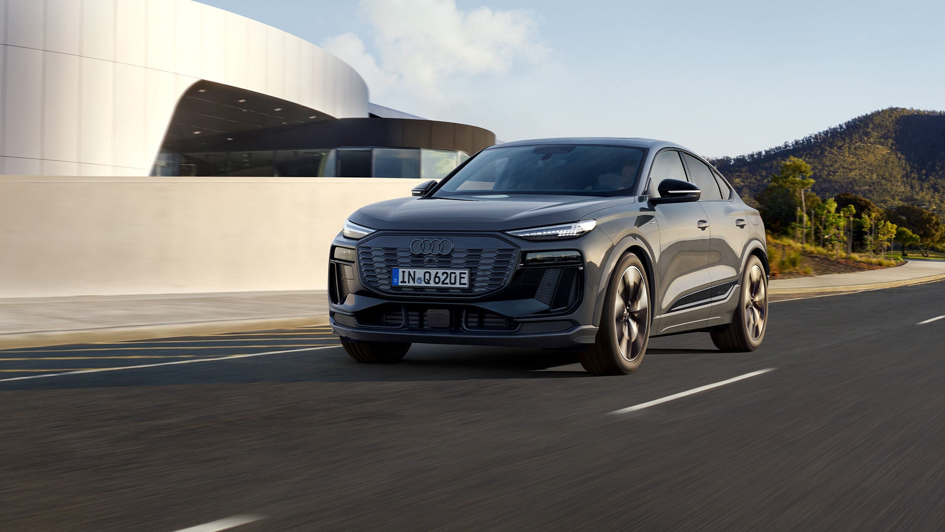 Grauer Audi Q6 Sportback e-tron fährt auf Straße vor moderner Architektur