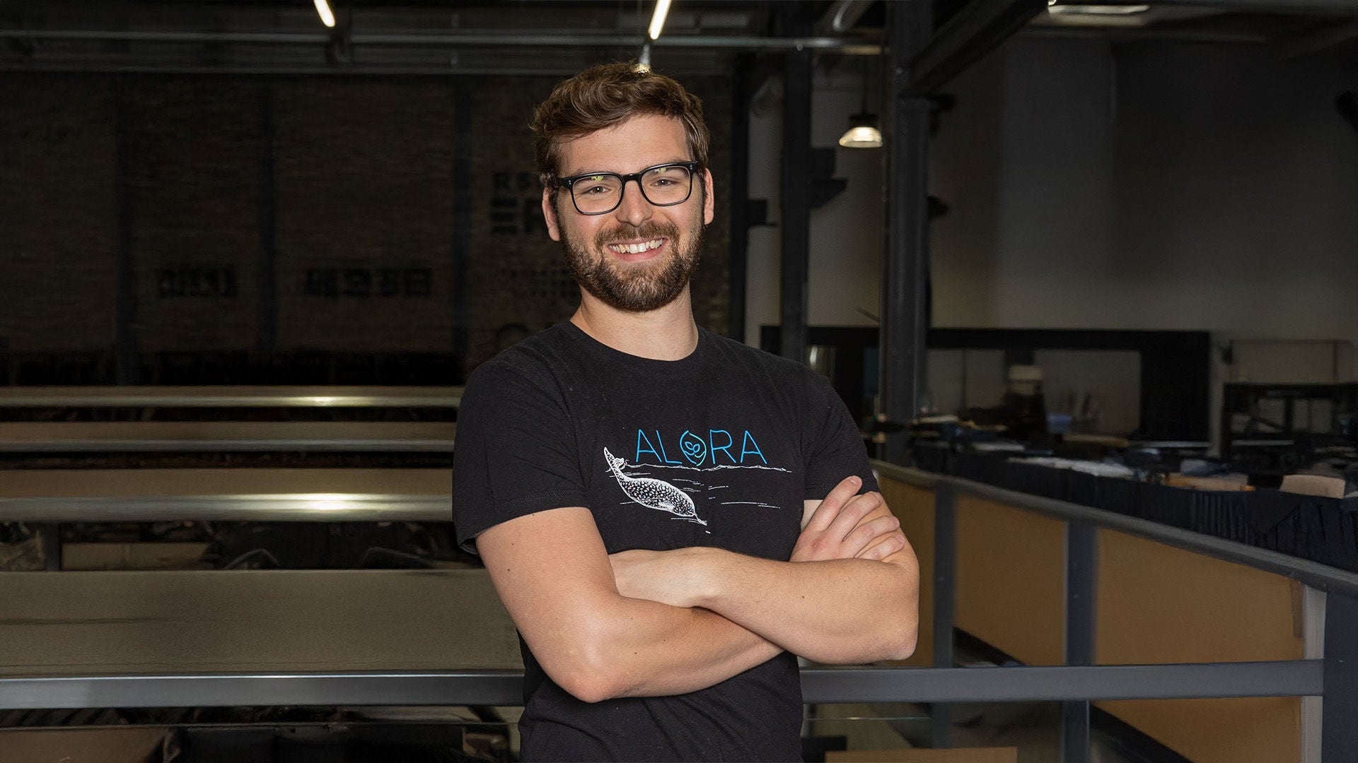 Mann mit Bart und Brille im schwarzen T-Shirt, Arme verschränkt, in einer Industriehalle stehend.