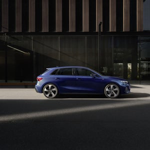 De Audi A3 Sportback, van achteren gezien schuin op een brug;  de ondergaande zon werpt een grote schaduw op de linkerkant van de auto.  Op de achtergrond is een kantoorgebouw zichtbaar.