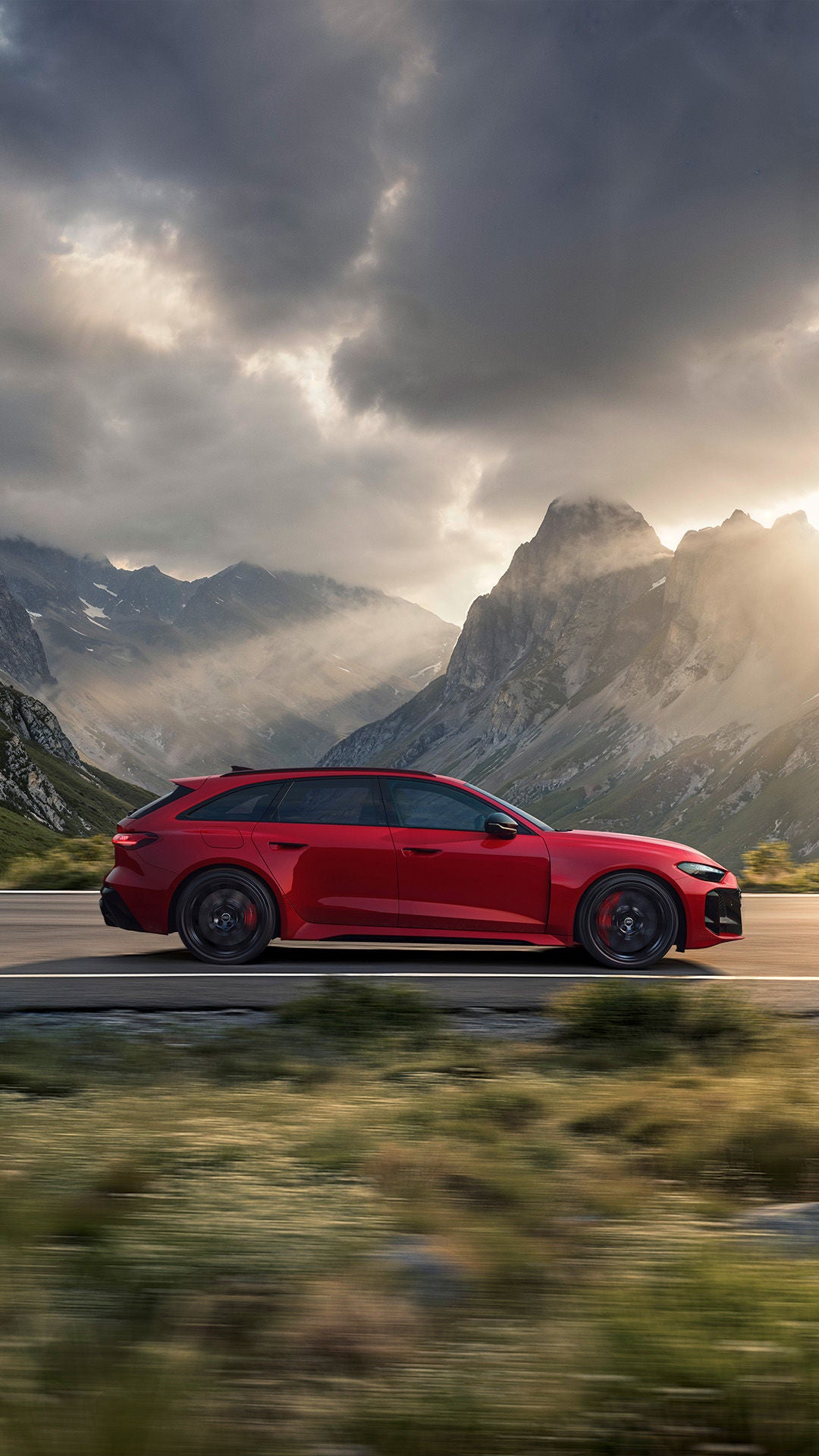 Red Audi RS 5 Avant station wagon driving on a mountain road, Alpine panorama with dramatic clouds and sunbeams in the background.