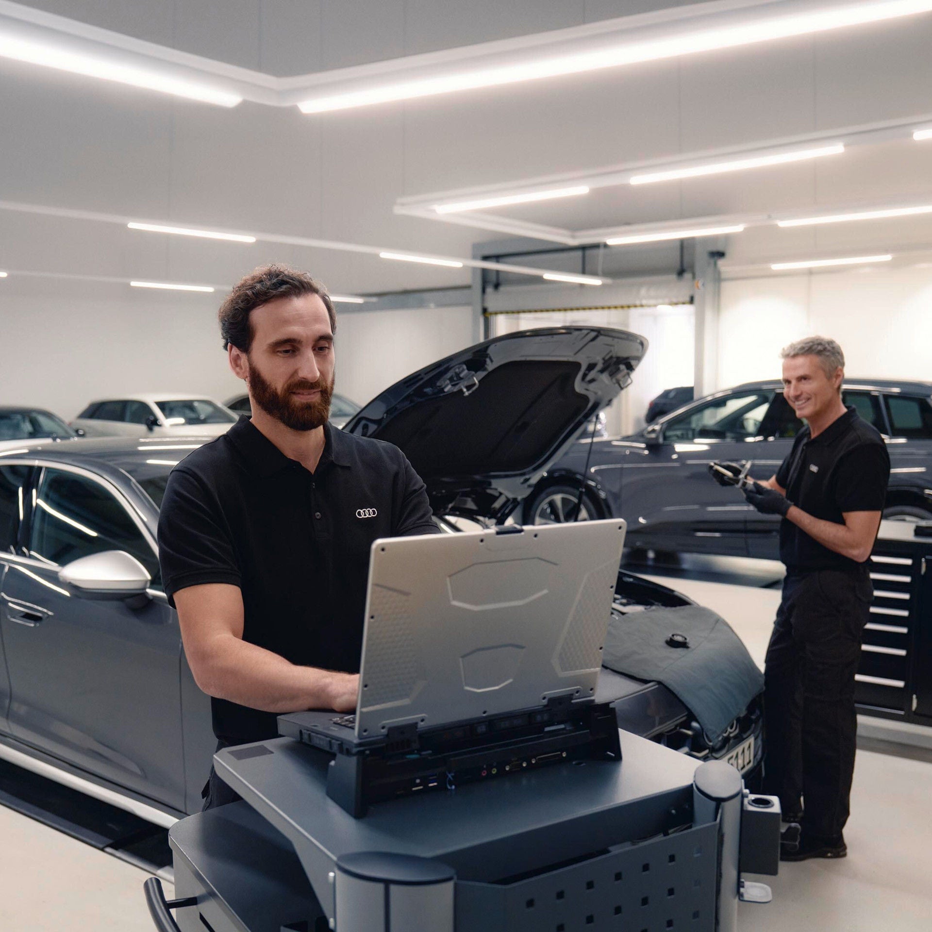 Zwei Mitarbeiter prüfen ein Auto mit geöffneter Motorhaube; einer arbeitet an einem Diagnosegerät.