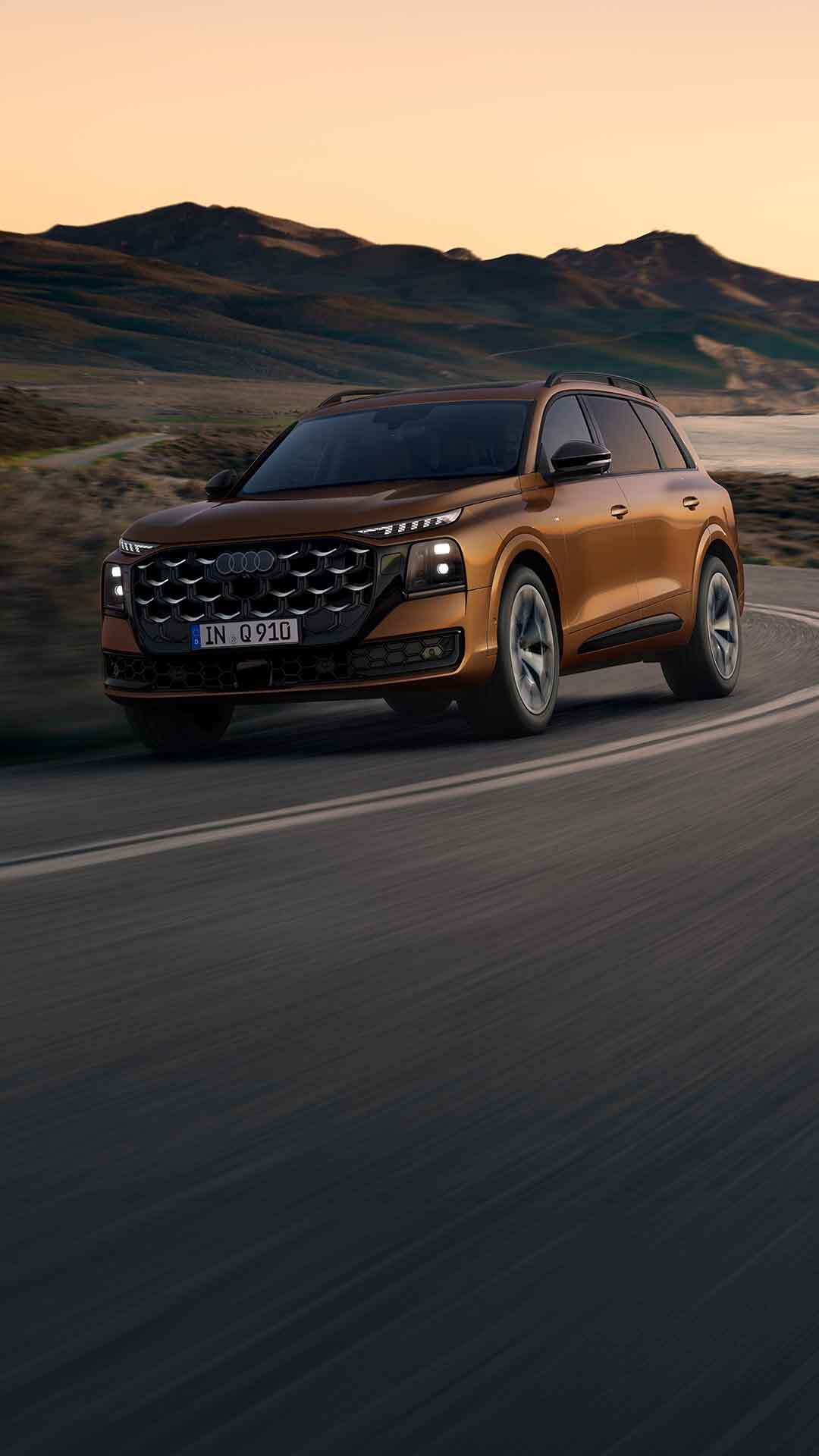 Der Audi Q9 SUV mit Audi exclusive Individuallackierung in Ipanemabraun Metallic, fotografiert schräg von vorn, durchquert eine Küstenstraße. Im Hintergrund das Meer und Gebirgszüge.
