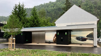 Überdachte Audi-Ladestation vor einem Haus mit Werbeplakaten, Bäumen und grüner Umgebung im Hintergrund.