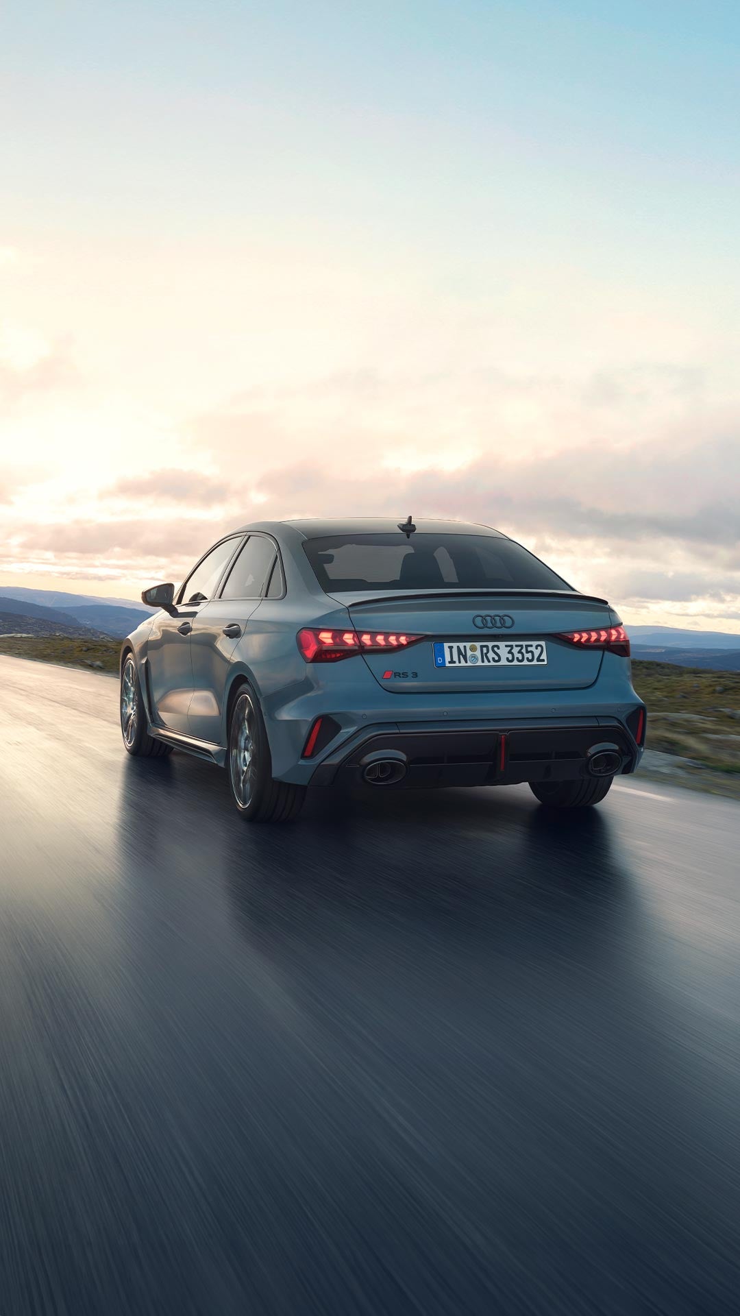 Audi RS 3 Sedan vista da dietro mentre percorre una strada di montagna sotto un cielo nuvoloso.