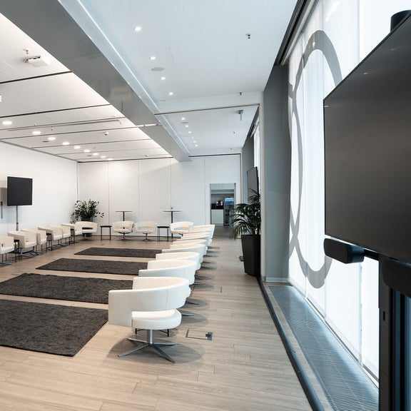 Heller Veranstaltungsraum mit weißen Sesseln in Reihen, Teppichen und großer Glasfront.