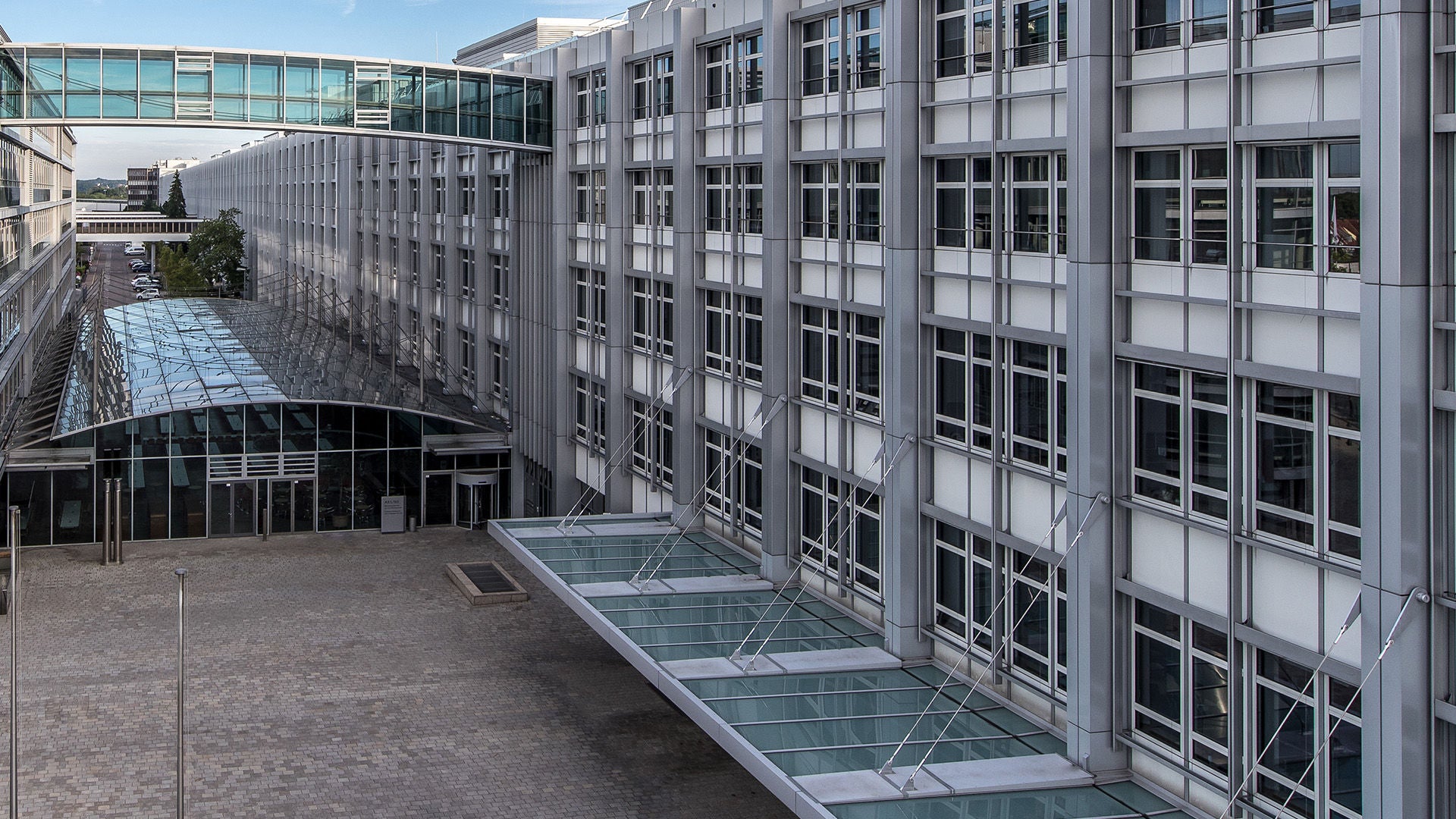 Modern office complex with glass facades and a glass bridge connecting two buildings, courtyard with paved plaza.