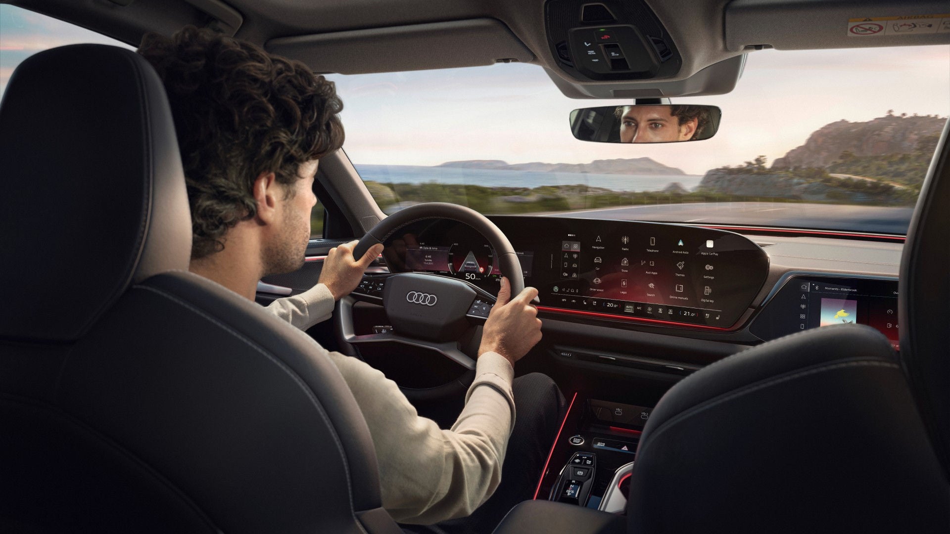 A man drives an Audi with a digital dashboard displaying navigation and controls. The view through the windshield shows a scenic, mountainous landscape.