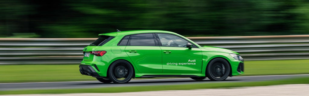 Light green Audi RS 3 Sportback in side profile on a race track; “Audi driving experience” lettering visible on the door.