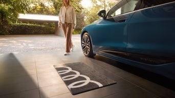 Teppich mit Audi-Ringen auf Fliesenboden neben einem parkenden Audi. Im Hintergrund läuft eine Frau entlang.