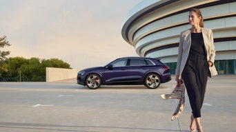 Frau in High Heels geht vor einem parkenden, blauen Audi Q8 SUV e-tron und modernem Gebäude entlang.