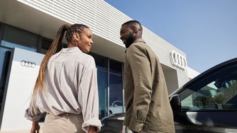 A man and woman walk smiling in front of an Audi dealership, with a glass façade and Audi logo in the background.