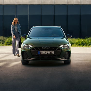 Frau steht neben einem grünen Audi A3 Sportback, frontal auf einem sonnigen Platz vor moderner Glasfassade.