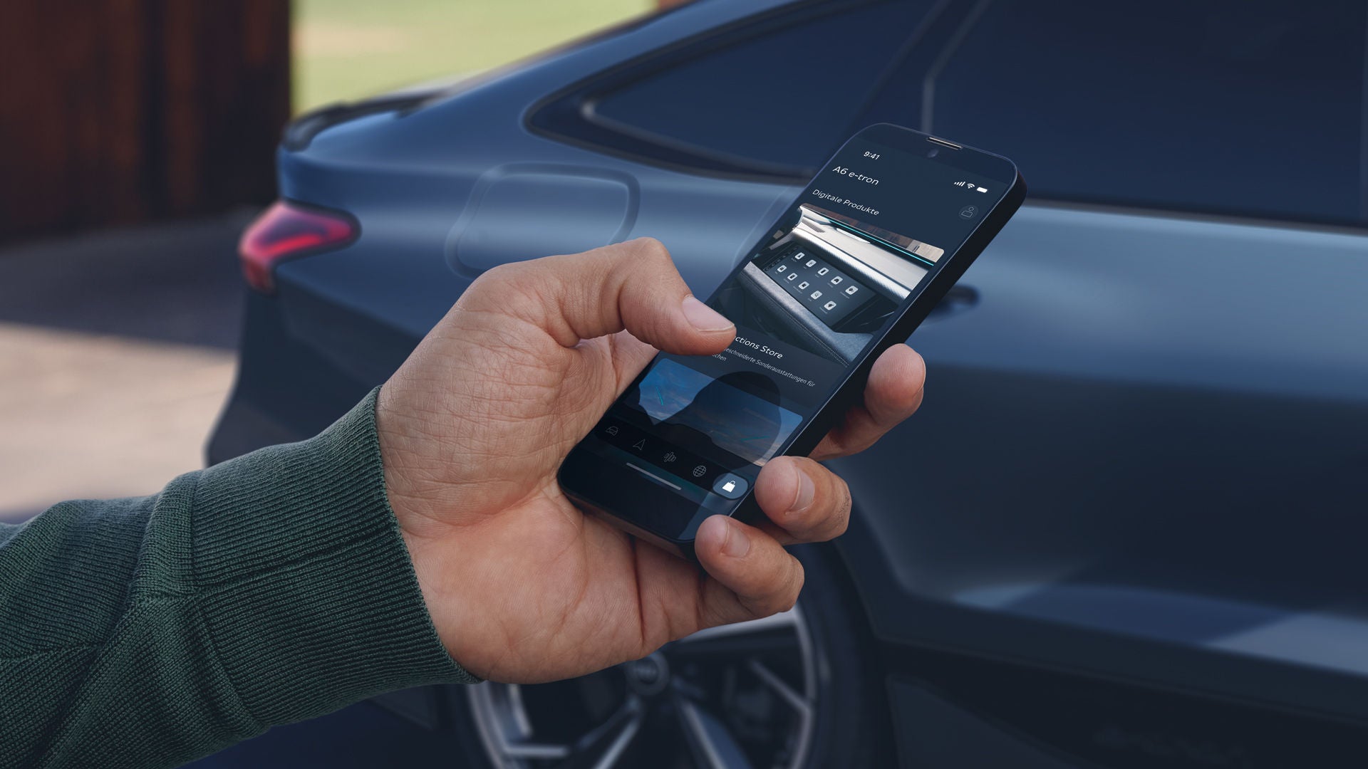 Die Hand eines Mannes ragt von links ins Bild. Er hält ein Smartphone in der Hand. Im Hintergrund steht leicht unscharf ein dunkelblauer Audi A6.