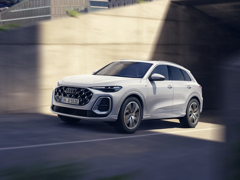 Angled, front view of a white Q5 SUV e-hybrid driving on a road in an urban environment.