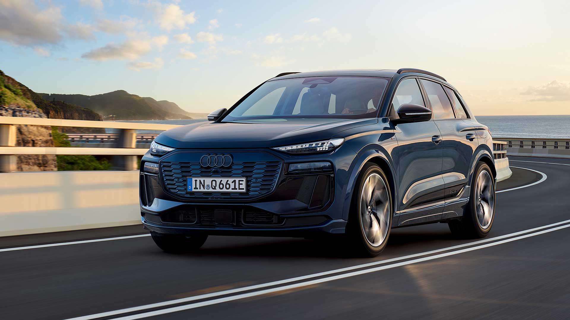Ein blauer Audi Q6 SUV e-tron performance fährt auf einer kurvigen Straße an der Küste entlang, bei Sonnenuntergang.