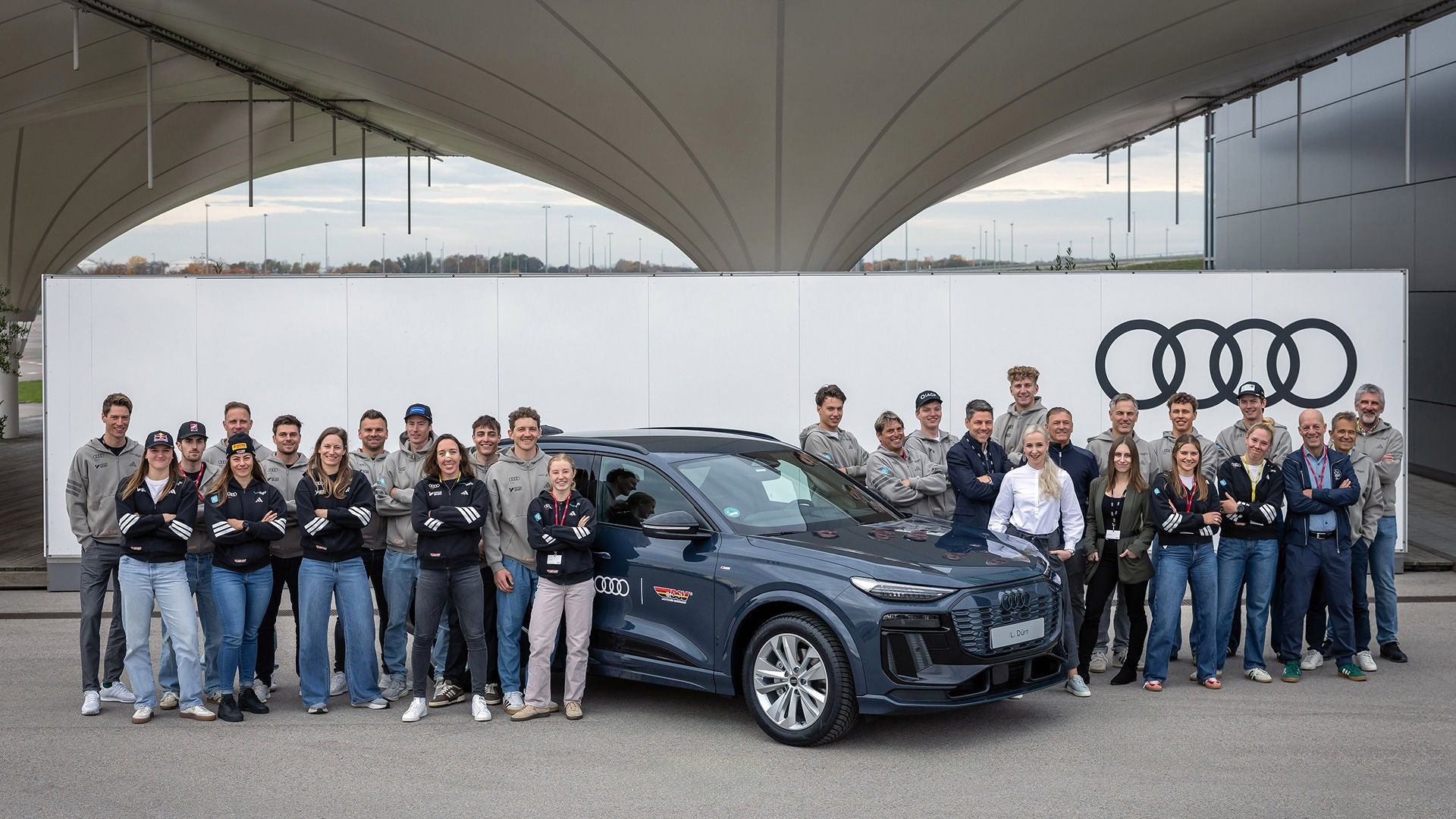 DSV-Top-Athletinnen und -Athleten am Audi Forum Ingolstadt vor einem grauen Audi Modell im Hintergrund sind rechts obern auf einer Wand die schwarzen Audi Ringe zu sehen.