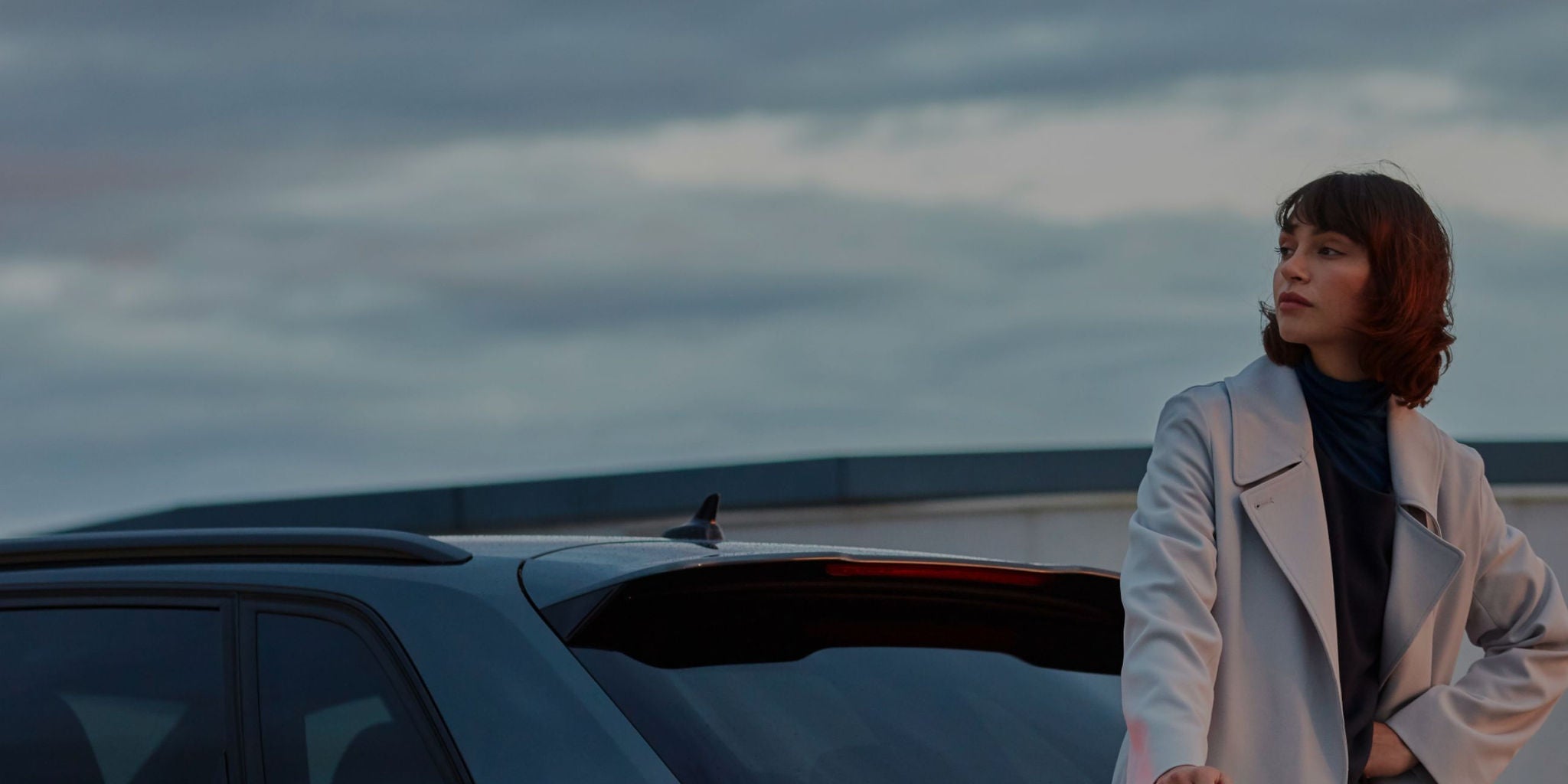 Person in a white coat standing next to a car with overcast skies in the background.