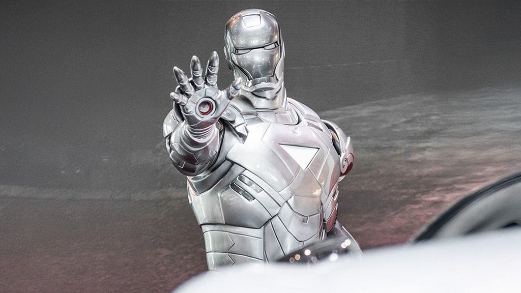 A silver Iron Man figure is positioned behind a white audi and reaching out with one hand.