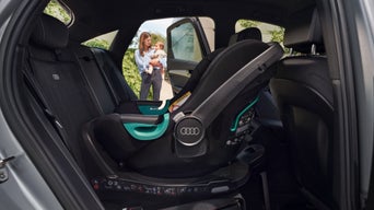 Babyschale auf Rückbank eines Audi, im Hintergrund Person mit Baby vor offenem Auto.