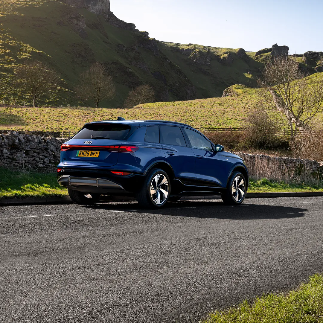 Blue Audi SUV parked on a quiet country road, shown from the rear three-quarter angle. Rolling green hills, rocky cliffs and leafless trees form the backdrop under clear daylight, highlighting the vehicle's sleek body lines, alloy wheels and continuous rear LED light strip.