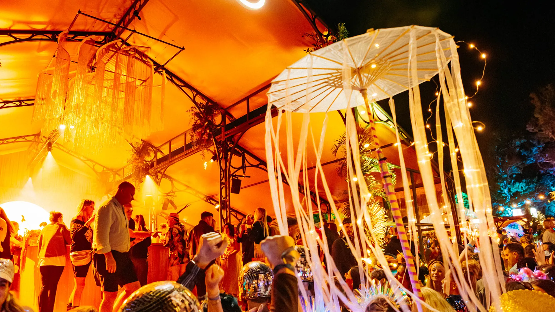 A glowing tasselled parasol hangs above a lively nighttime crowd, with golden chain chandeliers and string lights illuminating the tented area. A glowing tasselled parasol hangs above a lively nighttime crowd, with golden chain chandeliers and string lights illuminating the tented area.