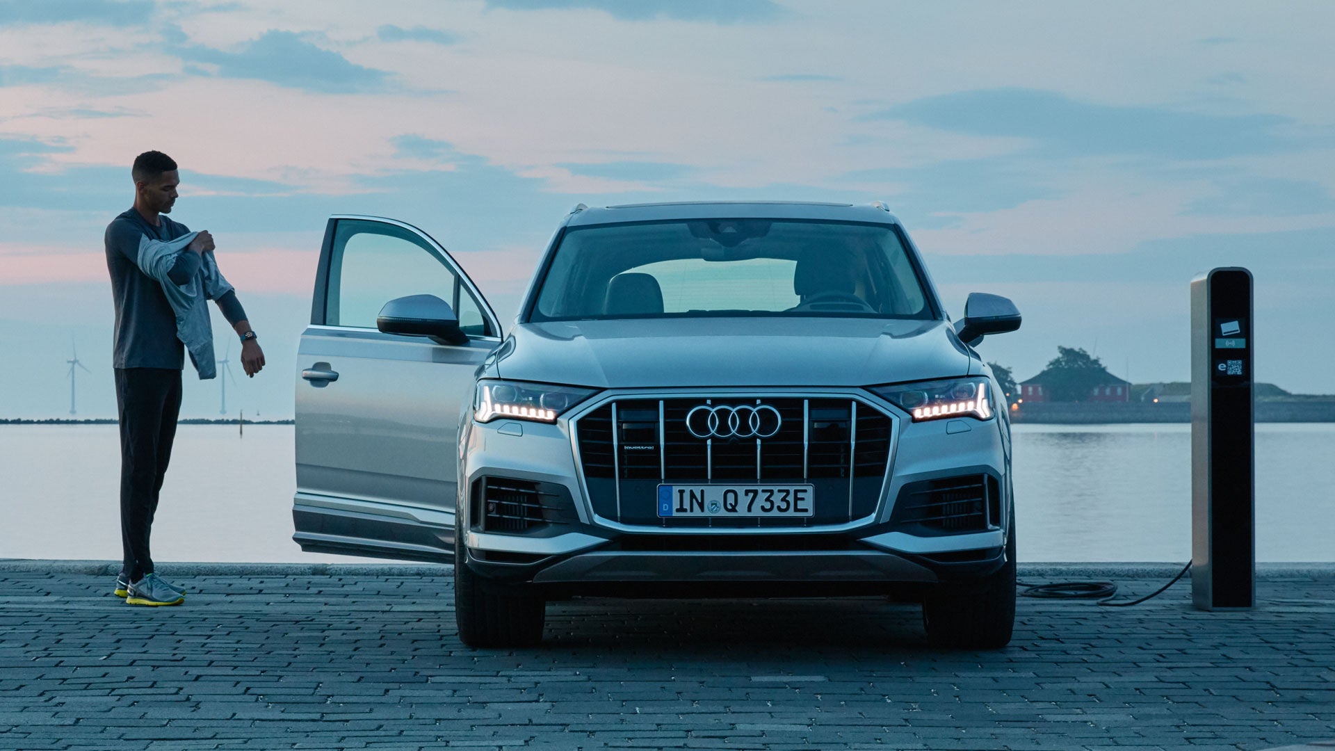 Audi Hybrid vor einem Gewässer an einer Ladesäule mit offener Tür, daneben steht ein Mann.