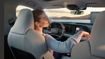 A woman relaxes in the passenger seat of a modern car with a digital dashboard overlooking a scenic waterfront at sunset, conveying a serene mood.
