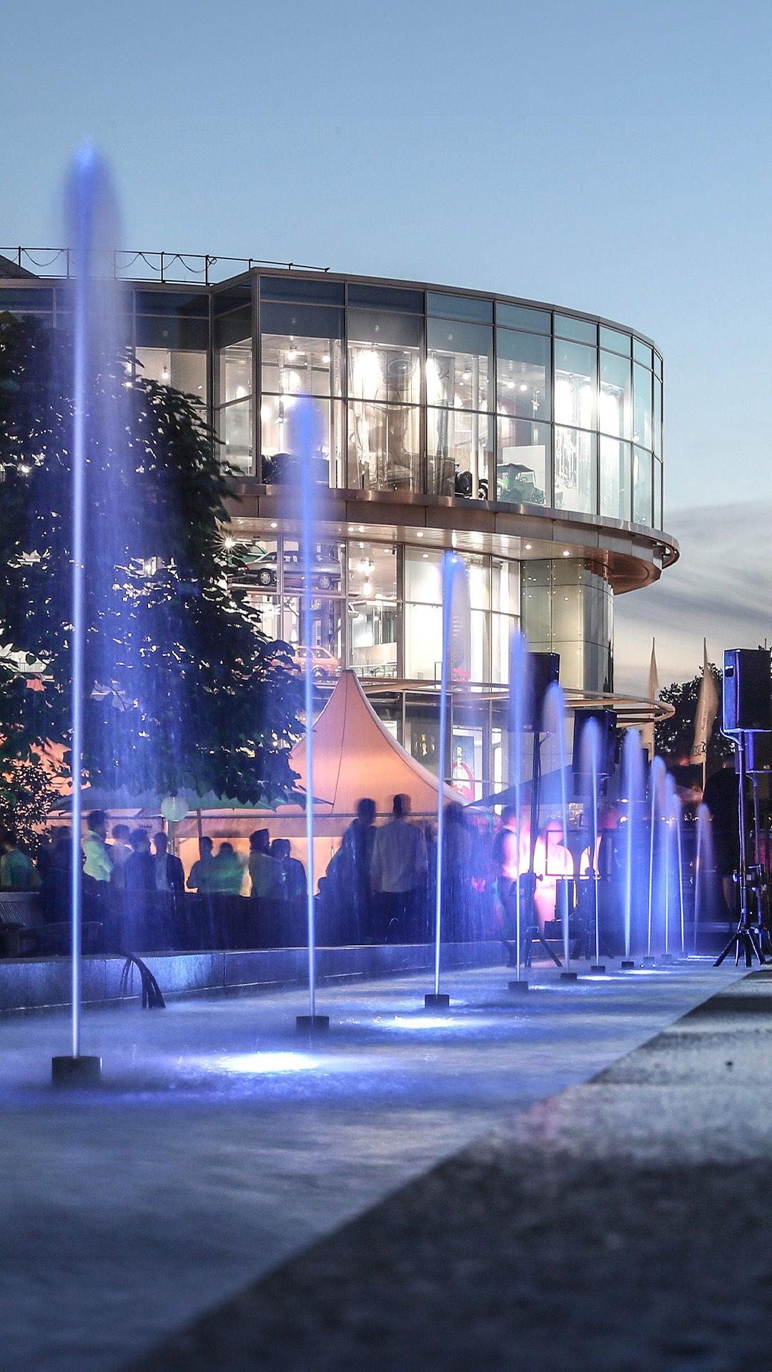 Modern glass façade of the Audi Forum at dusk, colorful light fountains and visitors in the foreground.