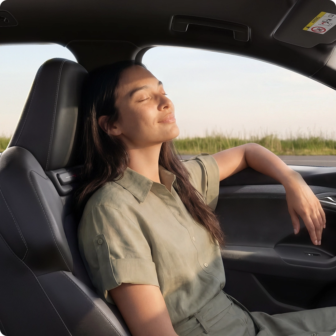 Interior of the Q6 SUV e-tron from the rear seat perspective. A young woman sits in the driver's seat and leans back with her eyes closed. Through the windscreen, you can see the sea and mountains.