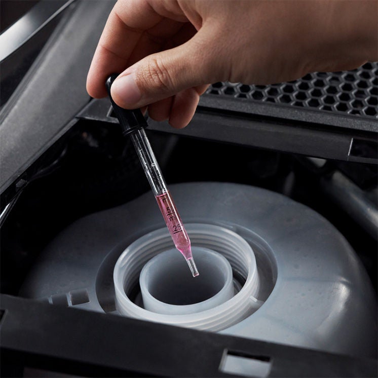 A hand holding a dropper that contains a pink fluid over an reservoir in an Audi engine