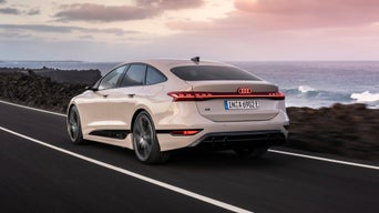 Audi sedan accelerates along a coastal road, showcasing its modern design against a backdrop of ocean waves and a colourful sky.