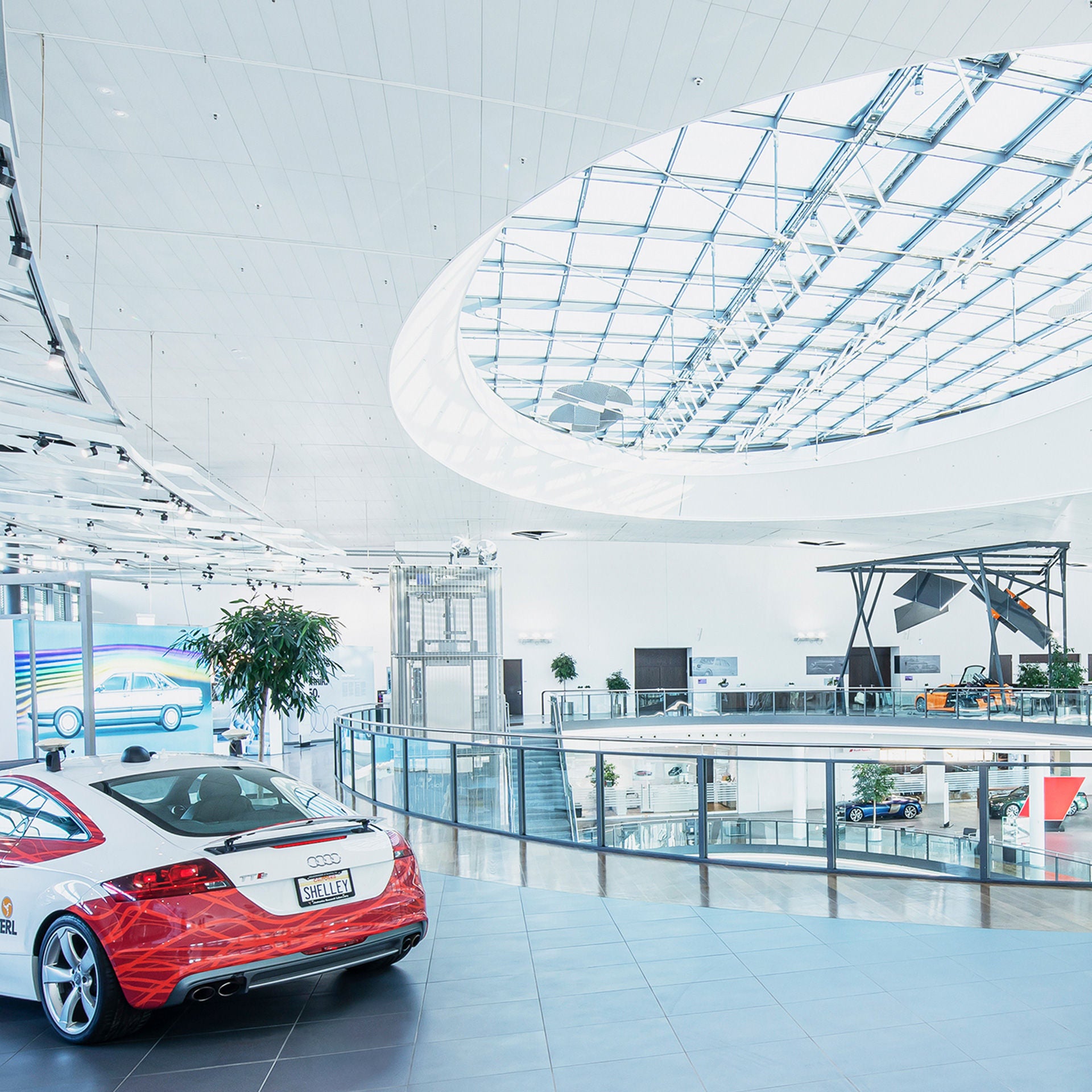 Helle moderne Autoausstellung mit mehreren Fahrzeugen, Glasdach, Galerie und Pflanze.