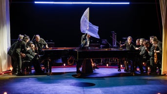 Musizierende auf einer blau beleuchteten Bühne. In der Mitte steht ein Mann am Klavier, der mit einer Hand eine weiße Fahne schwenkt.