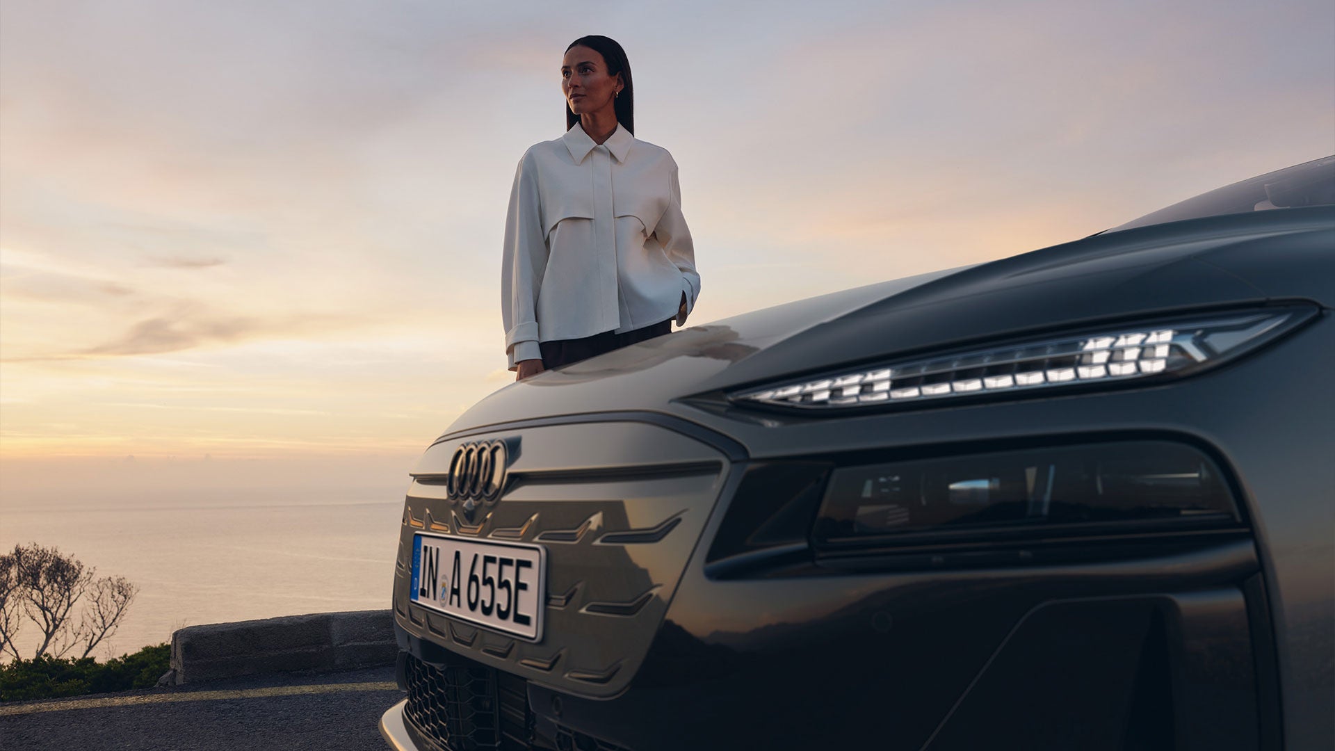 Frau steht bei Sonnenuntergang vor grauem Audi A6 Avant e-tron, Frontalansicht mit sichtbarem Kühlergrill.