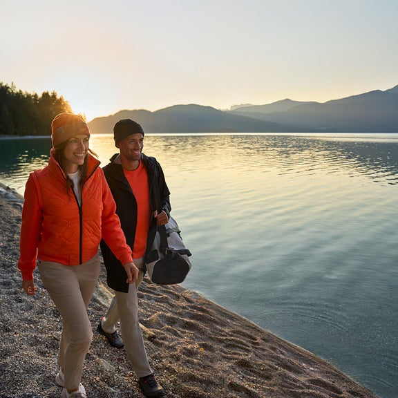 A couple walks along a lake at sunset. He is carrying a grey Audi sports bag.