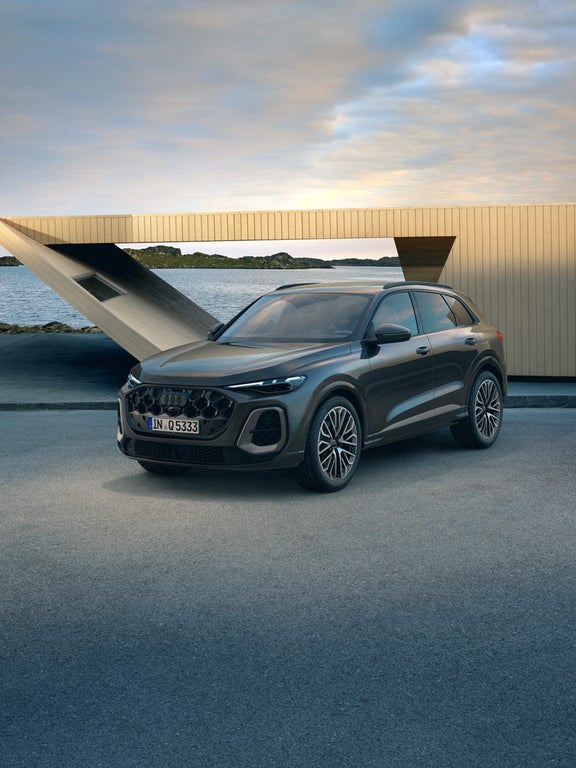 Ein grauer Audi Q5 SUV schräg von vorn vor einem modernen Holzgebäude an einer Wasserstraße.