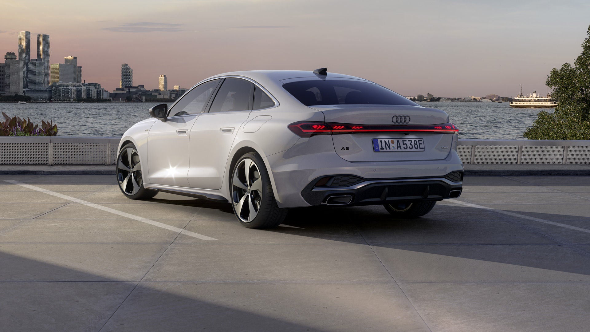 Die A5 Limousine e-hybrid steht im schrägen Heckprofil auf einem Parkplatz am Ufer eines Sees. Dahinter ist die Skyline einer Stadt bei Dämmerung zu sehen.