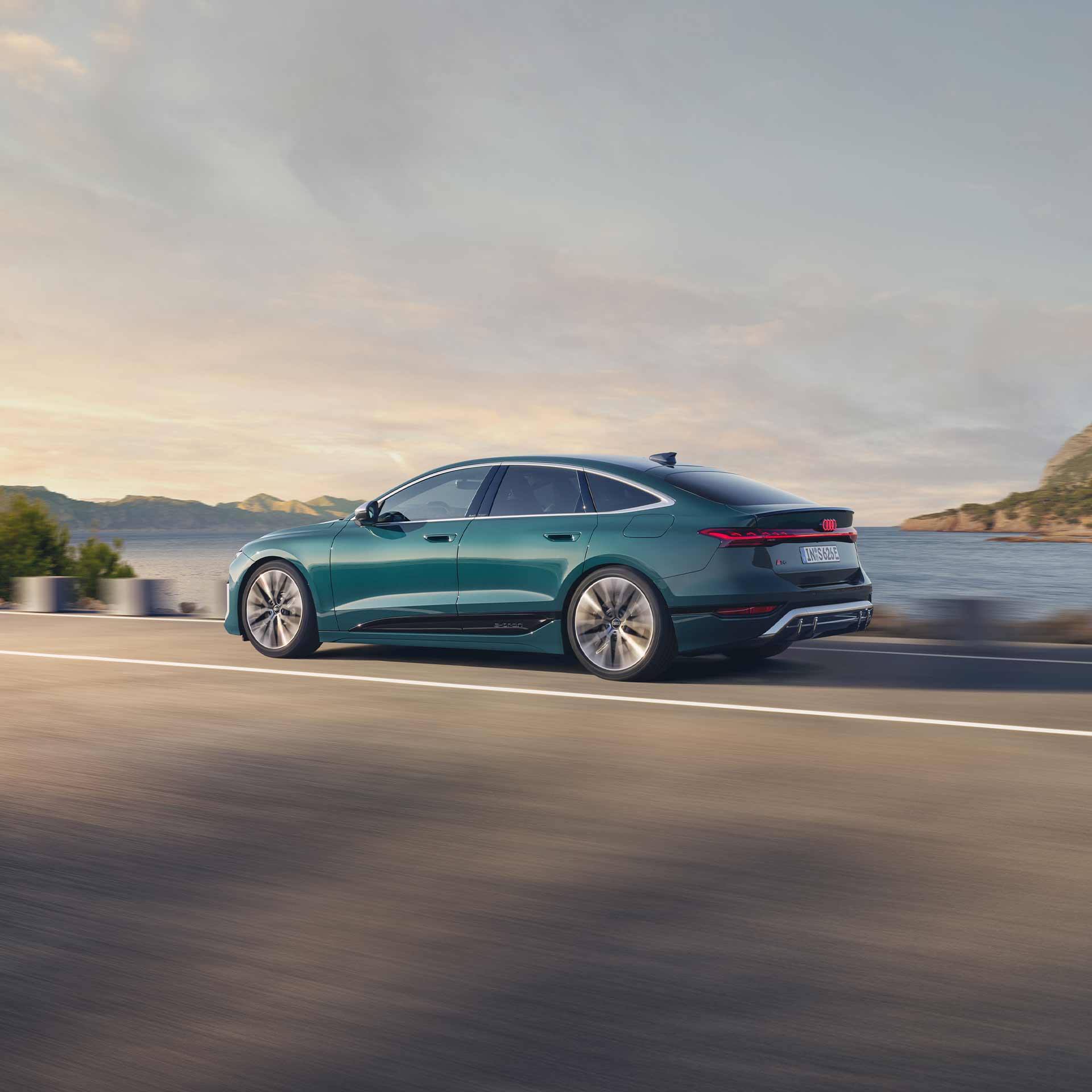Ein grüner Audi S6 Sportback e-tron fährt auf einer Küstenstraße, Meer und Hügel im Hintergrund.
