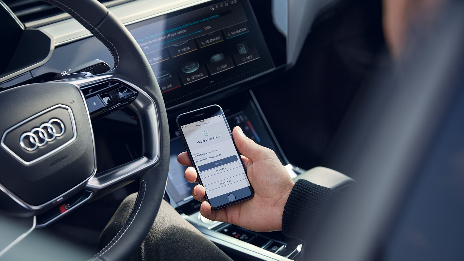 Ein Fahrer hält ein Smartphone mit einer Audi-App vor dem Lenkrad eines Audi-Fahrzeugs, im Hintergrund das zentrale Display.