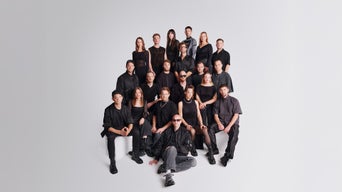 Große Musikgruppe in schwarzer Kleidung posiert im Studio vor hellem Hintergrund; mehrere Reihen, sitzend und stehend.