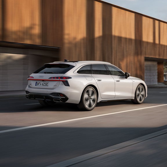Blick von schräg hinten auf den A6 Avant e-hybrid, der auf einer Straße vor einem Gebäude entlangfährt.