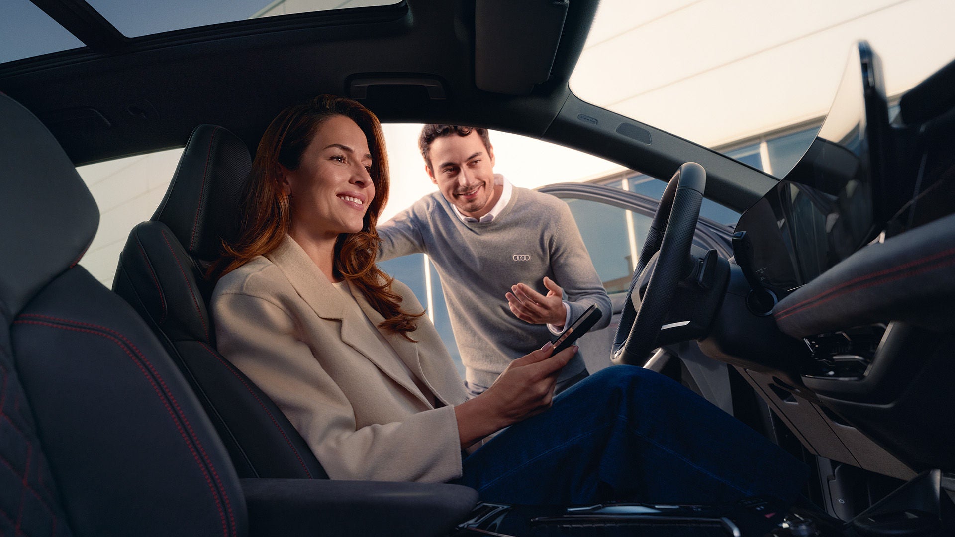 Frau sitzt lächelnd hinterm Steuer eines Audi Modell und hält ein Smartphone in der Hand, während ein Audi Mitarbeiter draußen am offenen Türrahmen steht und spricht.